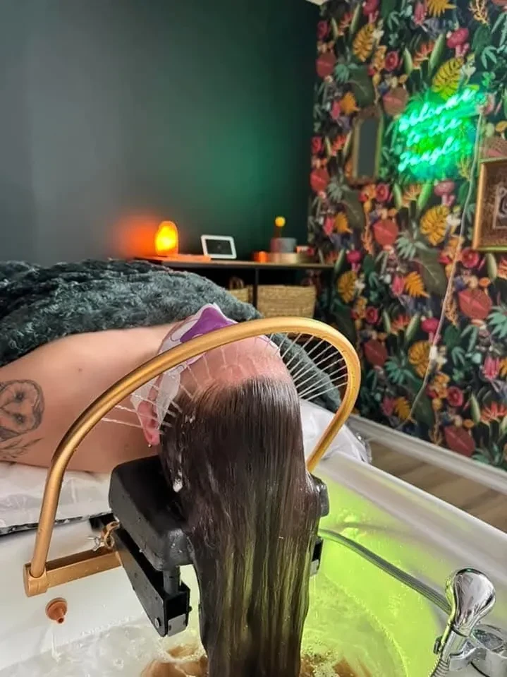 Client receiving a Japanese head spa treatment with water flowing over their hair at Ash's Lashes and Esthetics, with a cozy jungle-themed room in the background