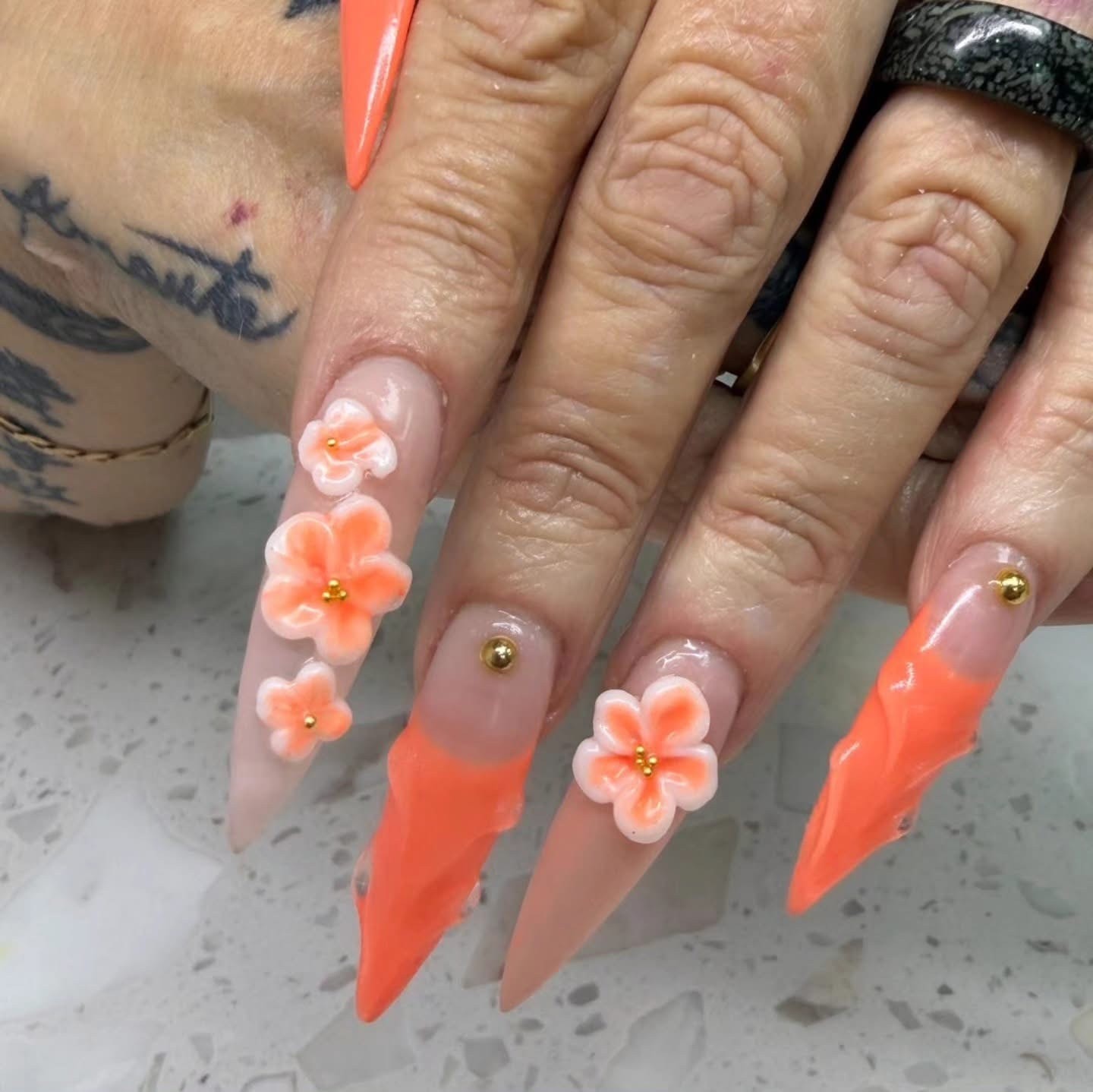 Coral orange stiletto nails with 3D flower embellishments and gold bead details at Ash's Lashes and Esthetics