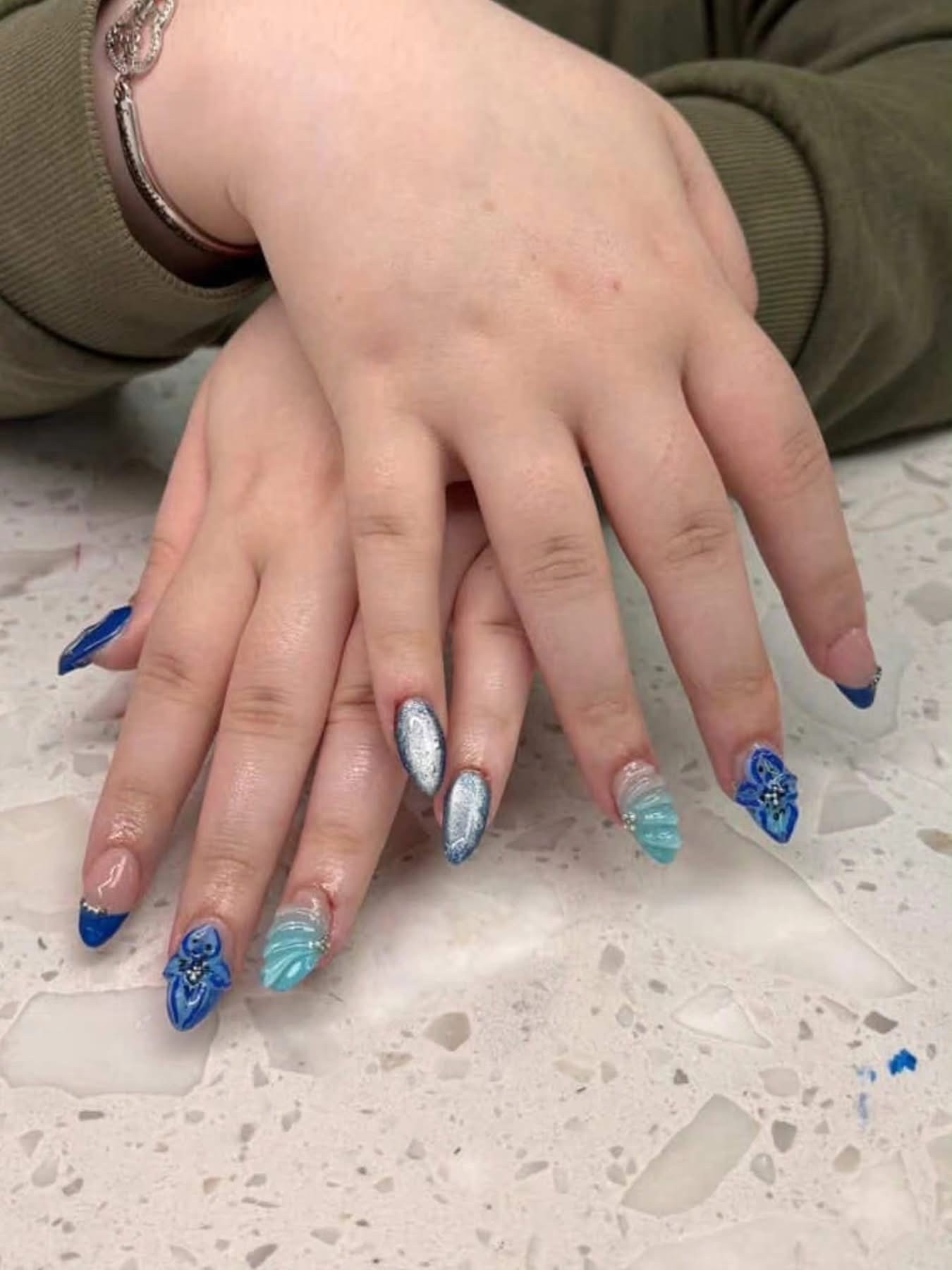 Blue and silver almond nails with 3D floral embellishments and glitter accent at Ash's Lashes and Esthetics