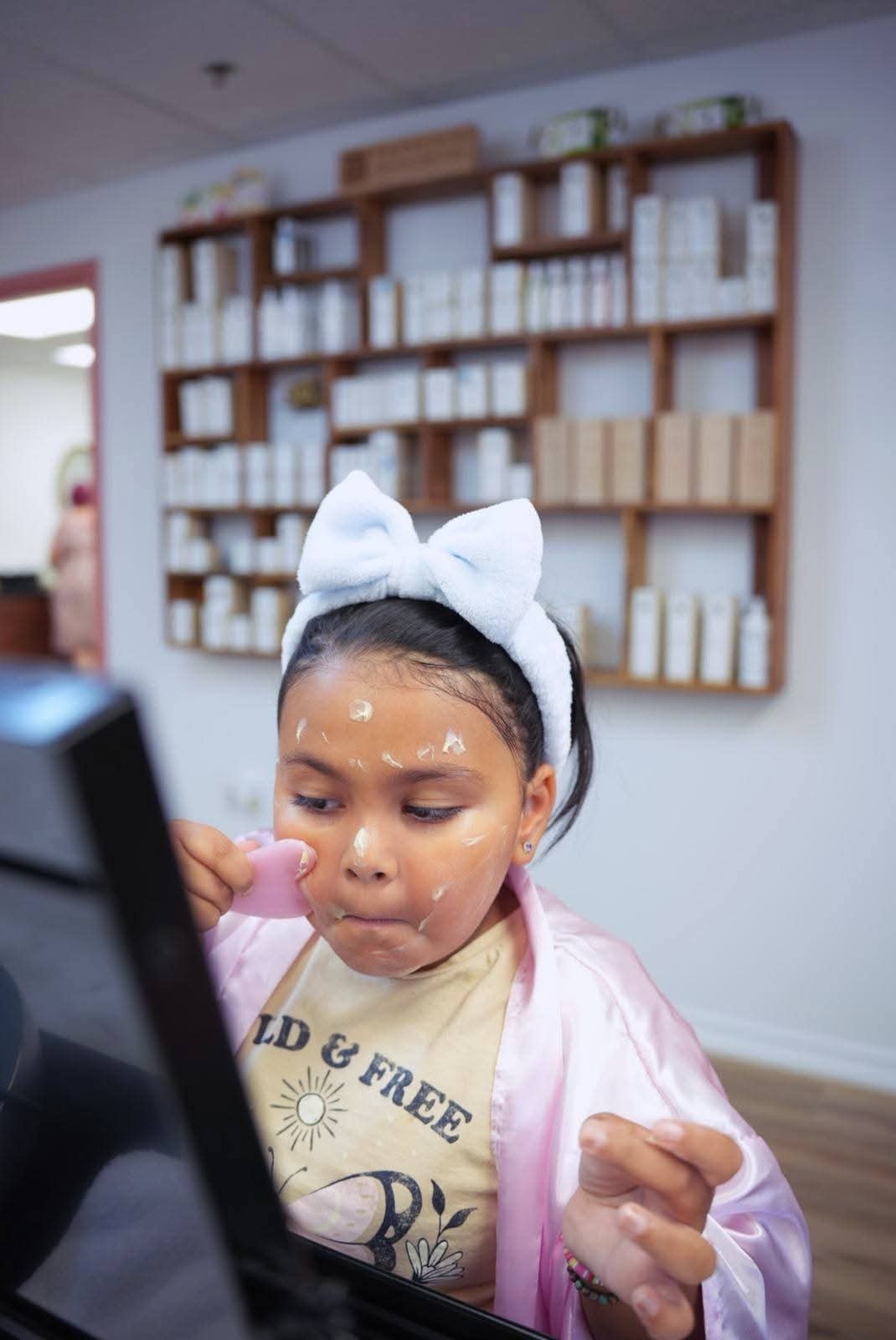 Young girl in a pink satin robe and bow headband using a gua sha tool on her face at Ash's Lashes birthday party