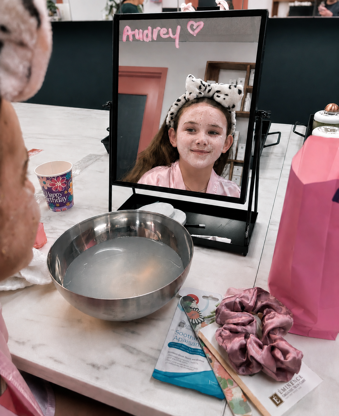 Birthday girl Audrey with a face mask and spa headband smiling in the mirror at Ash's Lashes