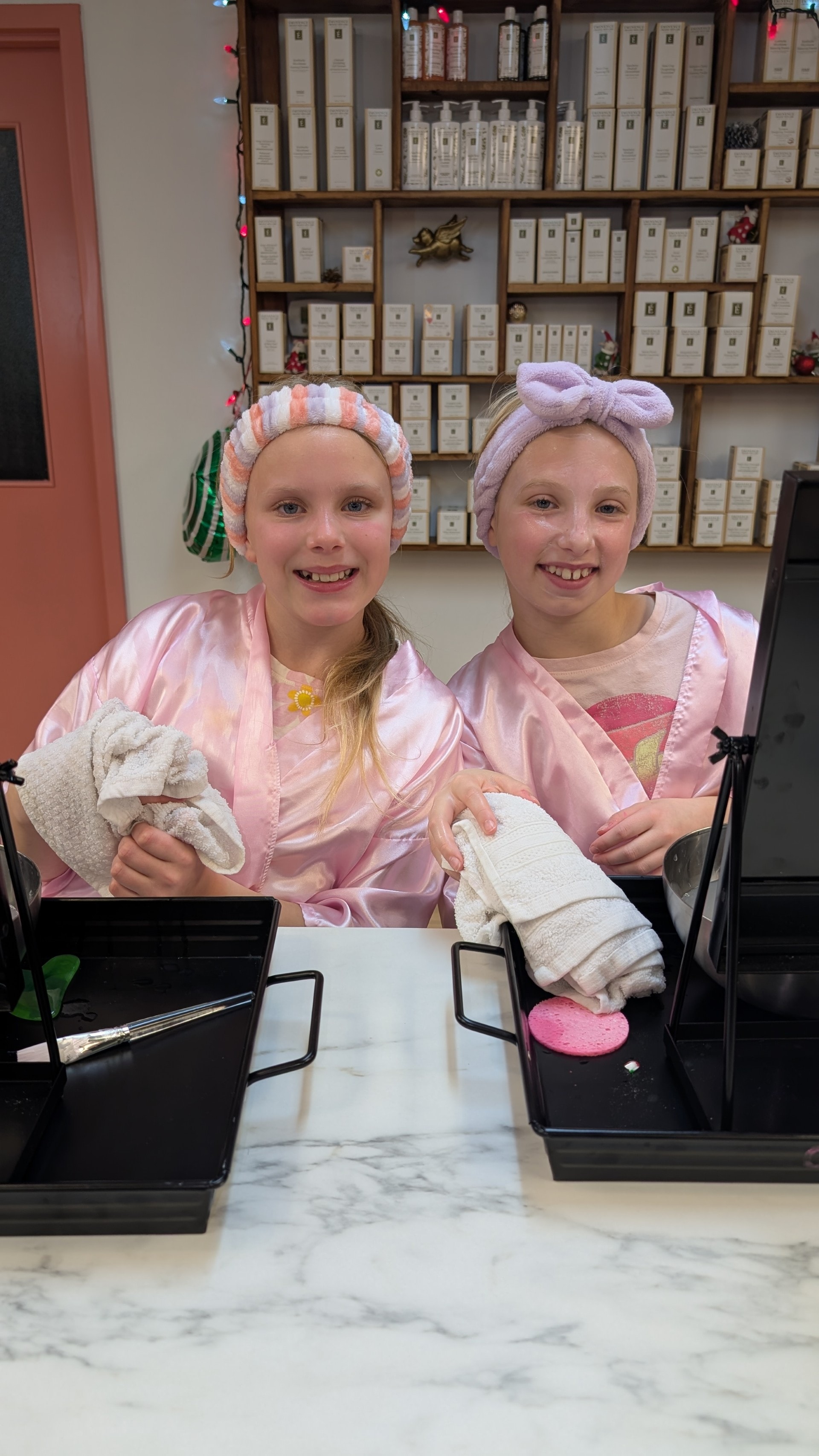 Two girls in pink satin robes and spa headbands smiling at the beauty station during a birthday party at Ash's Lashes
