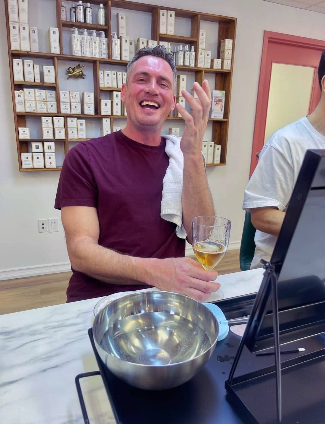 Man laughing while applying skincare with a towel and glass of wine during a You Dew You event at Ash's Lashes