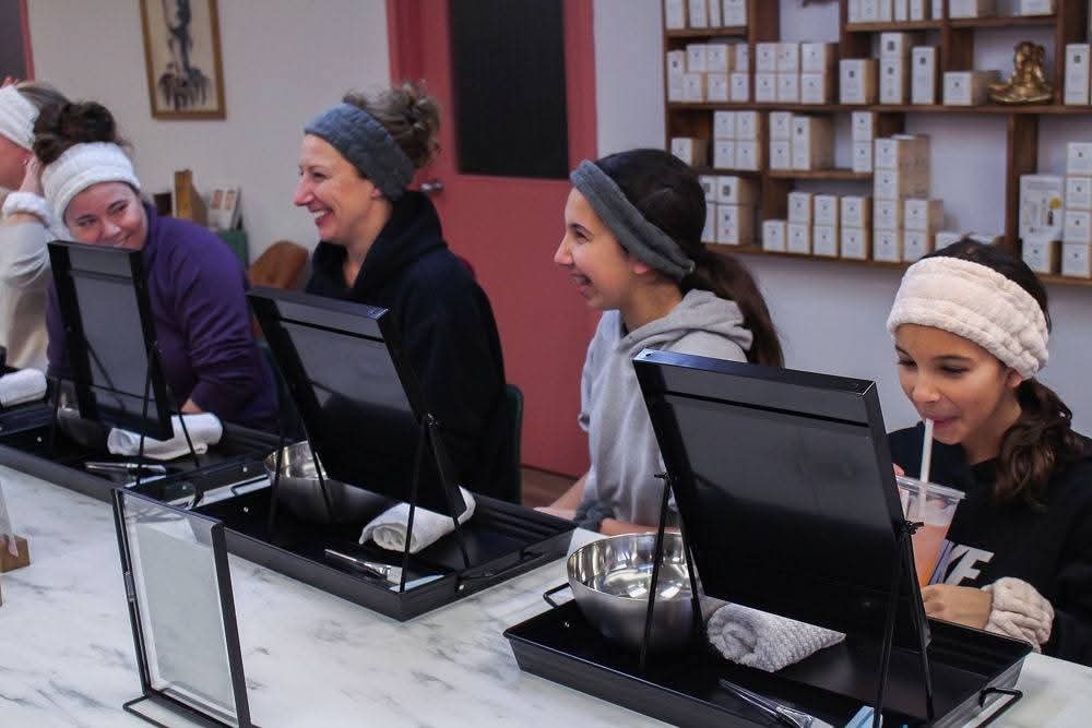 Group of women in spa headbands laughing and smiling at their skincare stations during a You Dew You guided facial event at Ash's Lashes